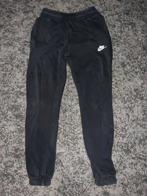 Svarta Nike mjukisbyxor - Svarta mjukisbyxor från Nike med vit logga på vänster lår. Byxorna har resår i midjan och vid bensluten samt sidofickor och en bakficka. Perfekta för chill eller träning, klassisk och enkel design.