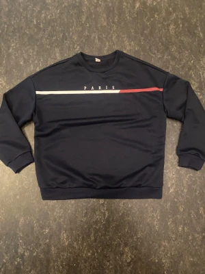 Mörkblå sweatshirt med PARIS-tryck - Snygg mörkblå sweatshirt med texten PARIS och en vit samt röd linje över bröstet. Tröjan har rund halsringning, långa ärmar och ribbade muddar vid ärmslut och nederkant. Perfekt för en avslappnad och trendig look.