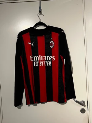 AC Milan långärmad matchtröja Puma - Säljer en långärmad AC Milan matchtröja från Puma med klassiska röda och svarta vertikala ränder. Tröjan har klubbmärke och Puma-logga på bröstet samt Emirates Fly Better-tryck på magen. Tillverkad i lätt och ventilerande material, perfekt för fotbollsträning eller match.