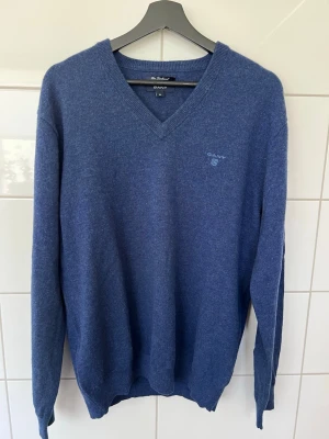 Blå stickad tröja från GANT - Blå stickad tröja från GANT i lammull. Tröjan har V-ringning, långa ärmar och diskret GANT-logga broderad på bröstet. Perfekt för dig som gillar klassisk och stilren design.