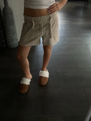 Beige och vita randiga shorts - Säljer ett par snygga shorts med beige och vita vertikala ränder. De har hög midja och en relaxed passform, perfekta för varma dagar. Shortsen har knäppning fram och är tillverkade i ett mjukt bomullsmaterial.