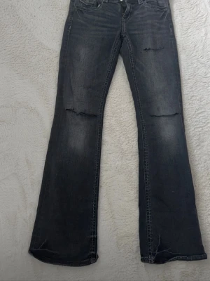 Mörkgrå bootcut jeans med slitningar - Säljer ett par mörkgrå bootcut jeans med slitna detaljer på låren och knäna. Jeansen har klassisk femficksmodell, normal midja och lätt utsvängda ben. Perfekta för en avslappnad och trendig look.  Går inte o köpa längre + egna hål 