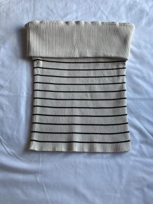 Randig ribbad tubtopp i beige och svart - Säljer en ribbad tubtopp i beige med svarta horisontella ränder. Toppen är axelbandslös och har en bred, vikt kant upptill för extra stöd. Perfekt för dig som gillar en enkel men trendig stil.