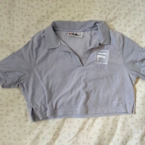 Ljusblå croppad t-shirt från FILA - Säljer en ljusblå croppad t-shirt från FILA med piké krage och korta ärmar. T-shirten har en broderad FILA-logga på bröstet och är gjord i mjuk bomull. Perfekt för en avslappnad och sportig stil.