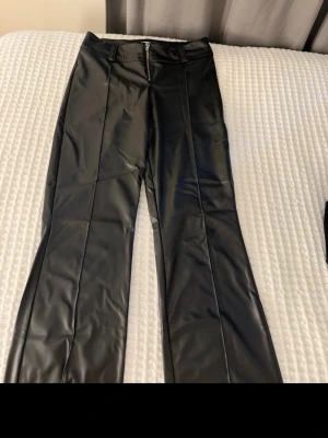 Svarta byxor i skinnimitation - Säljer ett par svarta byxor i skinnimitation med raka ben och hög midja. Byxorna har bälteshällor och dragkedja framtill. Snyggt glansigt material som ger en cool look.