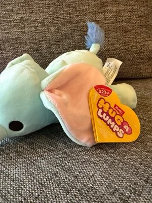 NY! Zuru Hug A Lumps 0,23kg Elefant Eliza - Mjukdjur i form av en elefant från ZURU Hug a Lumps. Färgen är ljusblå med rosa insida på öronen och en blå tofs på svansen. Ögonen är svarta och runda. Etiketten sitter kvar på mjukdjuret.  Ny!!