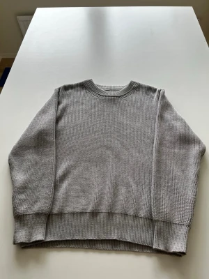 Grå oversized stickad tröja - En stilren grå stickad tröja med oversized passform och rund hals. Tröjan har breda ribbade muddar vid ärmslut och nederkant samt en diskret sömdetalj framtill. Perfekt för lager på lager och enkel att matcha. Passar i 60-65kg.