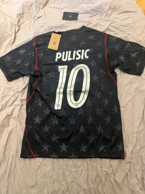 USA tröja Pulisic 10  - Säljer en svart Nike fotbollströja med stjärnmönster, röda detaljer och nummer 10 samt Pulisic på ryggen. USA-märke på bröstet och klassisk Nike-logga. Perfekt för fotbollsfans som vill sticka ut på planen.