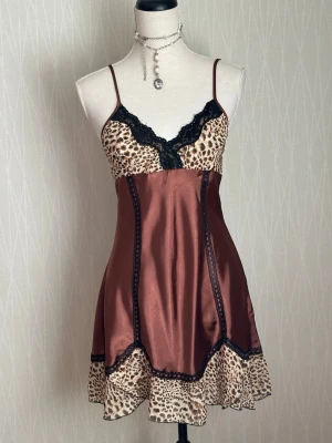 Brunt satin nattlinne med leopardmönster - Snyggt nattlinne i brunt satin med leopardmönstrade detaljer upptill och nertill. Tunna axelband och svart spets vid bysten ger en extra touch. Materialet är glansigt och mjukt, perfekt för en lyxig känsla hemma. Figurnära passform och kort modell.