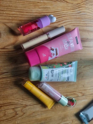 Blandat kit med skönhetsprodukter - Ett kit med olika skönhetsprodukter, inklusive ansiktsmask, cleanser, läppbalsam, concealer, handkräm och läppglans. Färgerna är rosa, lila, gul och mintgrön. Produkterna har olika former och lock, vissa med illustrationer och fruktdetaljer.