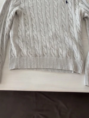 Ljusgrå kabelstickad tröja från  Ralph Lauren - Ljusgrå kabelstickad tröja med rund hals och långa ärmar. Tröjan har ribbade muddar vid ärmslut och nederkant samt en liten broderad logga på bröstet. Klassisk och tidlös design i mjukt material.