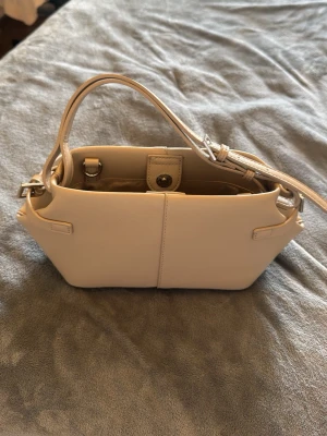 Beige handväska i stilren design - Snygg beige handväska i slät konstläder med unika vinklade kanter och justerbar axelrem. Väskan har ett huvudfack med tryckknappsstängning och silverfärgade detaljer. Perfekt för dig som gillar minimalistisk och modern stil. 