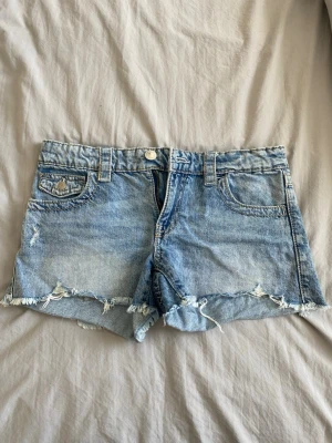 Ljusblå denimshorts med fransig kant - Säljer ett par ljusblå denimshorts med fransig, rå kant och slitna detaljer. Shortsen har fem fickor, bälteshällor och knappstängning fram. Perfekta för varma dagar och en avslappnad stil.