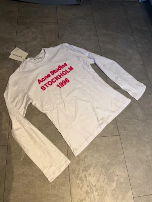 Vit långärmad tröja Acne Studios S - Super fin Acne Tröja. Vit långärmad topp från Acne Studios med rund hals och röd fluffig text på bröstet där det står 'Acne Studios STOCKHOLM 1996'. Tillverkad i mjukt bomullsmaterial och har en clean, minimalistisk look.
