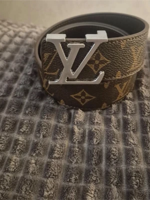 Brunt monogram skärp - Säljer ett brunt skärp med klassiskt monogrammönster och stor silvrig logga som spänne. Skärpet är gjort i präglat skinn och har guldfärgade detaljer i mönstret. Perfekt accessoar för att lyfta din outfit.