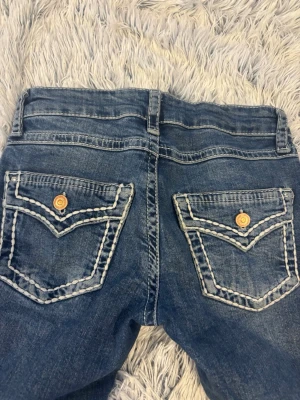 Blå jeans med kontrastsömmar - Snygga blå jeans med raka ben och tydliga vita kontrastsömmar på bakfickorna. Jeansen har klassisk femficksdesign och kopparfärgade knappar. Materialet är denim i bomull, perfekt för en avslappnad och trendig look.