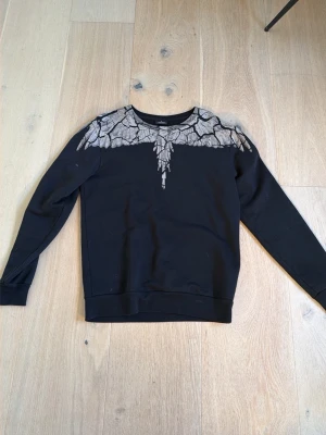 Svart sweatshirt Marcelo Burlon - Säljer en svart sweatshirt från Marcelo Burlon med unikt sprucket mönster i grått över axlar och bröst. Tröjan har rund halsringning och ribbade muddar vid ärmslut och nederkant. Tröjans krage är hyfsat stor. Perfekt för dig som gillar streetwear och coola detaljer. 