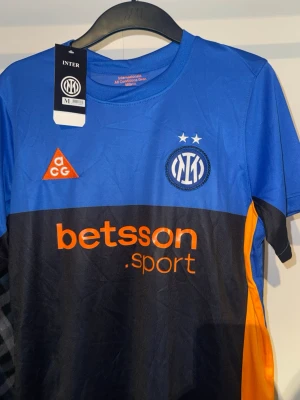 Blå Inter fotbollströja med tryck - Snygg blå och svart Inter fotbollströja med orange detaljer och tryck. Tröjan har klubbmärke, två stjärnor ovanför loggan och sponsortryck på bröstet. Tillverkad i ett lätt och funktionellt material som passar perfekt till träning eller match.