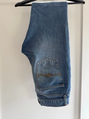 Blå raka jeans från Nudie Jeans - Säljer ett par klassiska blå jeans från Nudie Jeans med rak passform och snygga kontrastsömmar. Jeansen har en bakficka med den ikoniska Nudie-bågen och patch i läder bak i midjan. Tillverkade i slitstark denim av bomull.
