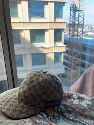 Gucci beige keps med GG-mönster - Säljer en beige keps från Gucci med det klassiska GG-mönstret över hela kepsen. På sidan finns en grön och röd rand som signaturdetalj. Kepsen har böjd skärm och justerbar rem bak. Perfekt för dig som gillar exklusiva accessoarer. Fler bilder kan givetvis ordnar:)