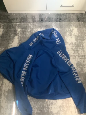 Blå hoodie med vit text - Säljer en blå hoodie med dragkedja och huva. Hoodien har vit text i gotisk stil längs båda ärmarna och på huvan. Praktiska fickor framtill och ribbade muddar vid ärmslut och nederkant. Perfekt för en avslappnad streetwear-look.