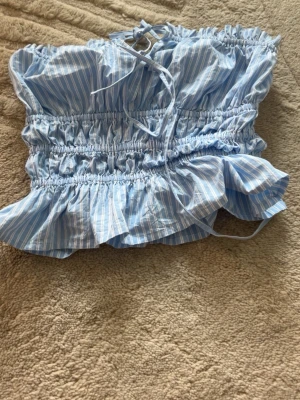 Blå- och vitrandig ärmlös topp - Supersöt blå- och vitrandig topp med volangkant och smockdetaljer. Toppen är ärmlös och har snörning upptill för justerbar passform. Perfekt för varma dagar och ger en fräsch och somrig vibe.