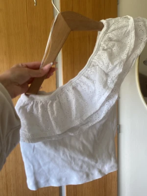 Vit oneshoulder topp med volang - Säljer en söt vit oneshoulder topp med bred volangkant och broderade detaljer. Toppen är ärmlös och har en feminin, somrig look som passar perfekt till jeans eller kjol.