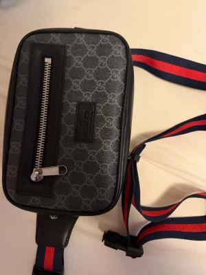 Svart magväska med mönster och röd-blå rem - Snygg svart magväska med grått monogrammönster och en framficka med dragkedja. Väskan har en justerbar rem i rött och blått, samt detaljer i svart konstläder. Perfekt för att bära det viktigaste och ger en trendig streetwear-look.