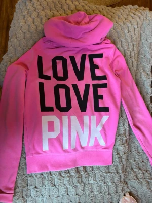 Rosa PINK hoodie med dragkedja och tryck - Säljer en rosa hoodie med huva och dragkedja. På ryggen står det 'LOVE LOVE PINK' i svart och vitt, och framtill finns en liten vit hundlogga. Hoodien har fickor fram och vita snören i huvan. Perfekt för en avslappnad och trendig look. Den är svår att få tag på så därifrån priset.