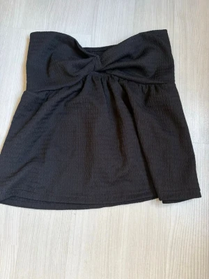 Svart bandeau tubtopp med twist - Säljer en svart bandeau tubtopp med twistad detalj framtill. Toppen är ärmlös och har en lätt strukturerad yta i mjukt material. Perfekt att styla med jeans eller kjol för en trendig look.