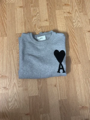 Grå stickad tröja med hjärtdetalj - Snygg grå stickad tröja från Ami med svart hjärta och bokstaven A broderat på bröstet. Tröjan har rund halsringning, ribbade muddar och nederkant. Perfekt för dig som gillar stilrena och trendiga plagg.