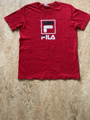 Röd t-shirt från FILA med logga - Snygg röd t-shirt från FILA med stor vit och svart logga på bröstet. Klassisk rak passform och rund halsringning. Tillverkad i mjukt bomullsmaterial som känns skönt mot huden. Perfekt för en sportig och avslappnad stil.