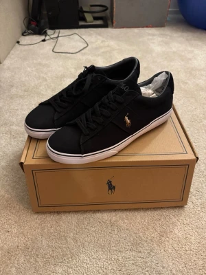 Svarta Polo Ralph Lauren sneakers - Stilrena svarta sneakers från Polo Ralph Lauren med klassisk låg profil och vit sula. Skorna har en guldfärgad logga på sidan och är tillverkade i textil med matchande svarta skosnören. Storleken är mer 46 1/3 - 47 än 46.⭐️