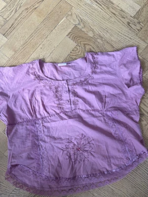 Ljuslila blus med spetsdetaljer - Söt ljuslila/rosa blus med korta ärmar och rund halsringning. Blusen har broderade blommor och spetsdetaljer framtill samt dekorativa knappar vid halsen. 🌸🤗