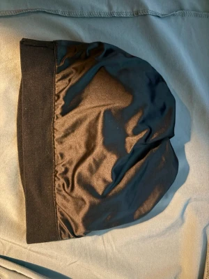 Svart mössa från Sats - Svart mössa med enkel design och Satyn-logga broderad på kanten. Utsidan är i mjukt tyg medan insidan har ett glansigt, slätt foder som känns skönt mot håret. Perfekt för dig som vill ha en stilren och bekväm mössa till vardags.