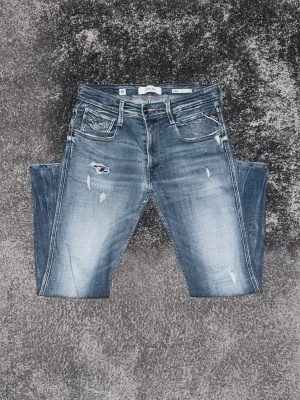 Blå slitna jeans med tvättade detaljer - Säljer ett par blå jeans med slitningar och tvättade detaljer. Jeansen har fem fickor, dragkedjegylf och en klassisk passform. Materialet är denim och färgen är mellanblå med ljusare partier och distressed look.