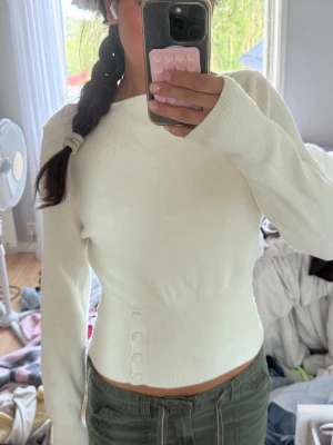 Vit tröja med knappar - Säljer en vit, långärmad tröja med rund hals och dekorativa knappar vid midjan. Tröjan har en figurnära passform och är tillverkad i ett mjukt, stretchigt material som sitter snyggt på kroppen.