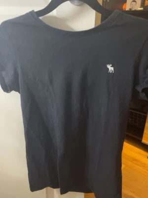 Marinblå t-shirt med liten logga - En enkel marinblå t-shirt i bomull med korta ärmar och rund hals. På bröstet finns en liten vit logga som ger en clean och stilren look. Perfekt till jeans eller shorts för en avslappnad stil.