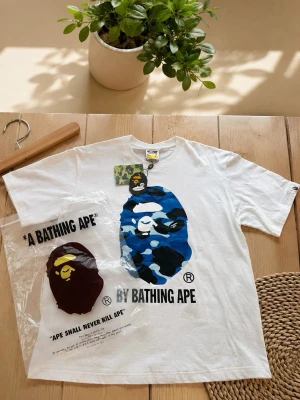 BAPE Color Camo Ape Tee vit/blå - Säljer en vit t-shirt från BAPE med stort färgglatt camo-tryck av apansikte i blått, grönt och vinrött på framsidan. Klassisk rund hals och korta ärmar. Ikonisk streetwear med tryckt logga och text. Perfekt för dig som gillar unik design.