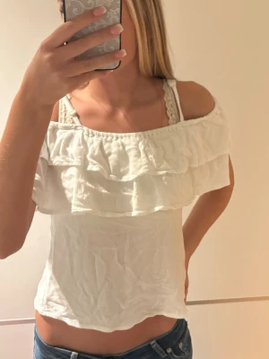 Vit offshoulder topp med volang - Söt vit offshoulder topp med dubbla volanglager upptill. Toppen är ärmlös och har en luftig, lätt passform som passar perfekt till sommaren. Tillverkad i ett mjukt bomullsliknande material och har en fräsch, ljus look.