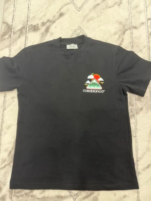 Svart t-shirt från Casablanca - Svart t-shirt från Casablanca med färgglad grafik på bröstet som visar ett berg, sol och moln. T-shirten har rund halsringning och korta ärmar. Tillverkad i mjuk bomull för en skön känsla.