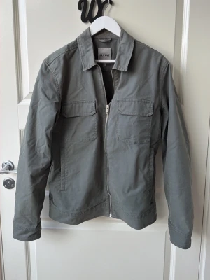 Jacka från Elvine i strl L - Snygg grå overshirt från Elvine med dragkedja framtill, två bröstfickor med lock och raka ärmar. Tillverkad i ett mjukt bomullstyg som känns bekvämt och stilrent. Perfekt lager-på-lager-plagg för en clean look.