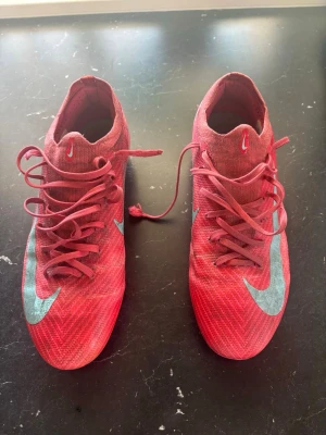 Röda Nike fotbollsskor med blå swoosh - Säljer ett par röda Nike fotbollsskor med blå swoosh på sidan. Skorna har snörning och låg profil, samt mönstrad ovandel i syntetmaterial för bra bollkontroll. Perfekta för dig som vill sticka ut på planen!
