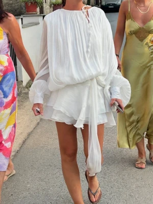 Vit sommarklänning med volang - Superfin vit klänning med omlottdesign och volangdetaljer. Klänningen har långa, vida ärmar med vita pärlknappar vid ärmslut och ett knytband i midjan. Det ingår även ett par shorts till som man har under klänningen, se sista bilden😍  Storleken är S/M