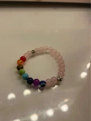 Färgglatt pärlarmband i rosa och regnbåge - Säljer ett elastiskt armband med ljusrosa pärlor och inslag av färgglada pärlor i regnbågens färger. Armbandet har även två silvriga detaljer som ger extra stil. Perfekt accessoar för dig som gillar färg och vill sticka ut.