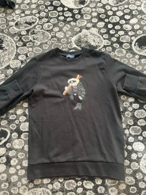 Svart sweatshirt med björntryck - Cool svart sweatshirt med ett broderat motiv av en björn i vinterkläder och skidutrustning på bröstet. Tröjan har rund halsringning och långa ärmar. Perfekt för dig som gillar streetwear och unika detaljer.