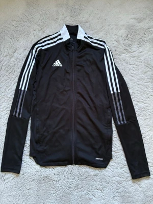 Svart Adidas träningsjacka  - Säljer en svart träningsjacka från Adidas med vita ränder längs ärmarna och vit detalj på kragen. Jackan har hel dragkedja framtill, ståkrage och är tillverkad i Primegreen-material. Perfekt för träning eller uppvärmning.