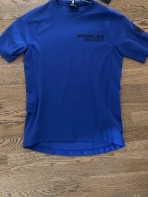 Blå t-shirt med tryck - Snygg blå t-shirt med svart tryck på bröstet där det står '#MONCLER DAY-NAMIC'. T-shirten har rund hals och korta ärmar. Perfekt för en stilren och sportig look.