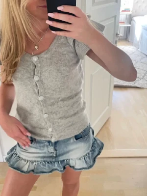 Ljusgrå stickad topp med knappar - Supersöt ljusgrå stickad topp med korta ärmar och stora vita knappar framtill. Toppen har en rundad halsringning och ribbade kanter. Super mjuk & skön🤍