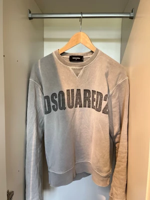 Grå sweatshirt från DSQUARED2 - Säljer en grå sweatshirt från DSQUARED2 med stor svart logga över bröstet. Tröjan har rund halsringning, ribbade muddar vid ärmslut och nederkant samt en avslappnad passform. Perfekt för en casual och trendig look.